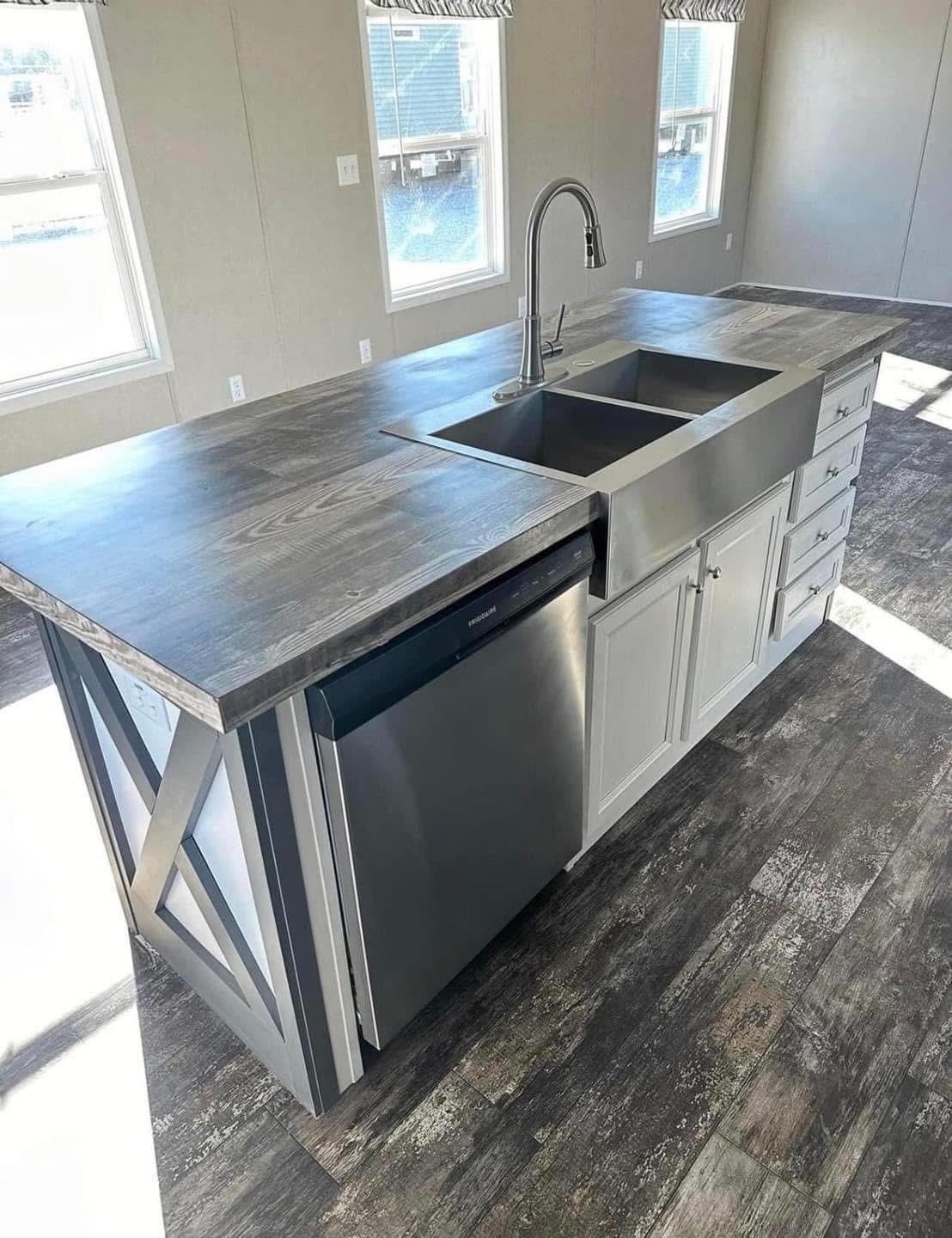 Modern kitchen island with a stainless steel sink, faucet, and dishwasher. It features a wooden countertop and gray cabinets, in a sunlit room.