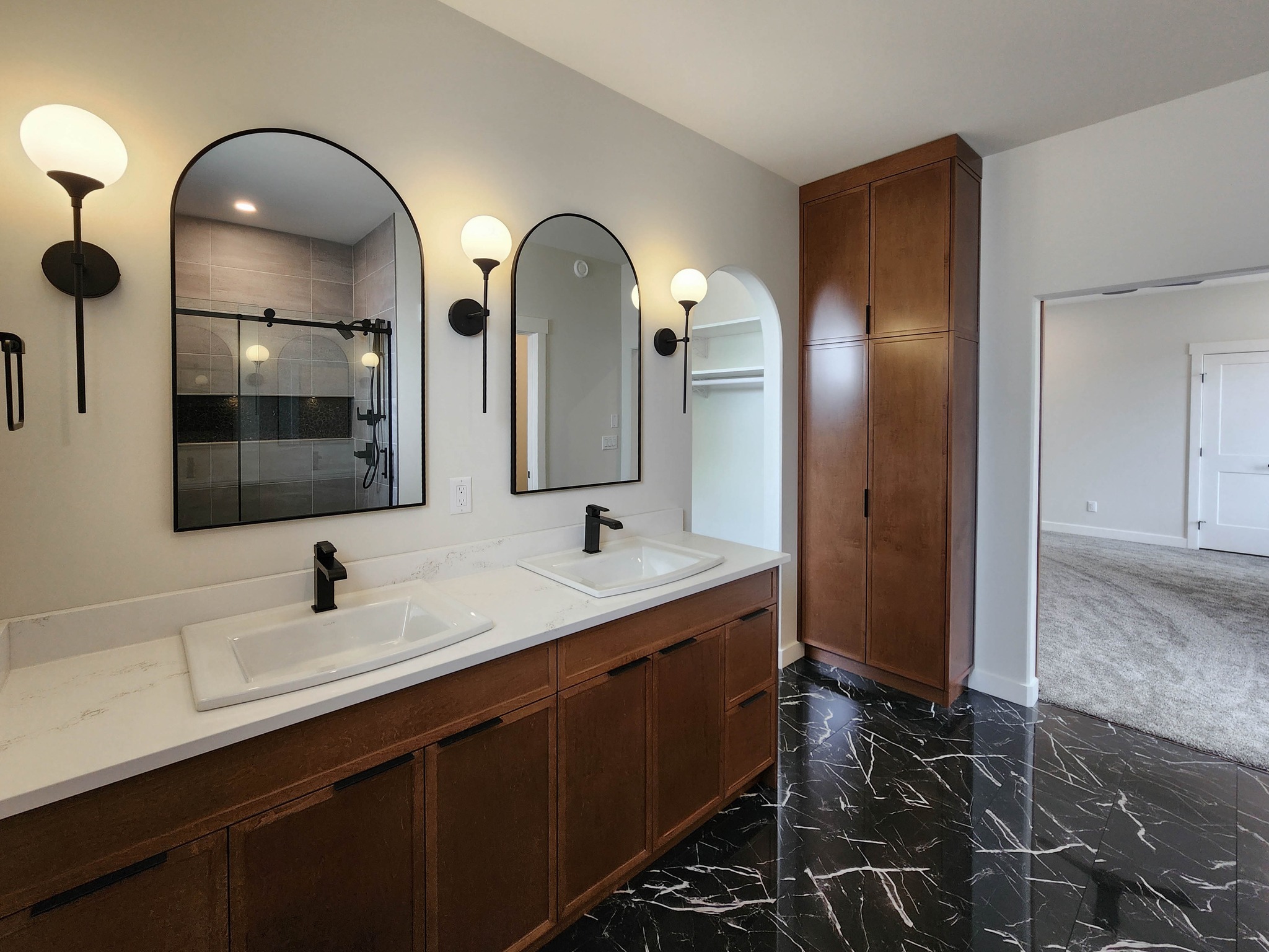 Modern bathroom with dual sinks, marble floor, and wood cabinetry. Two arched mirrors reflect a walk-in shower. Soft lighting creates a serene ambiance.