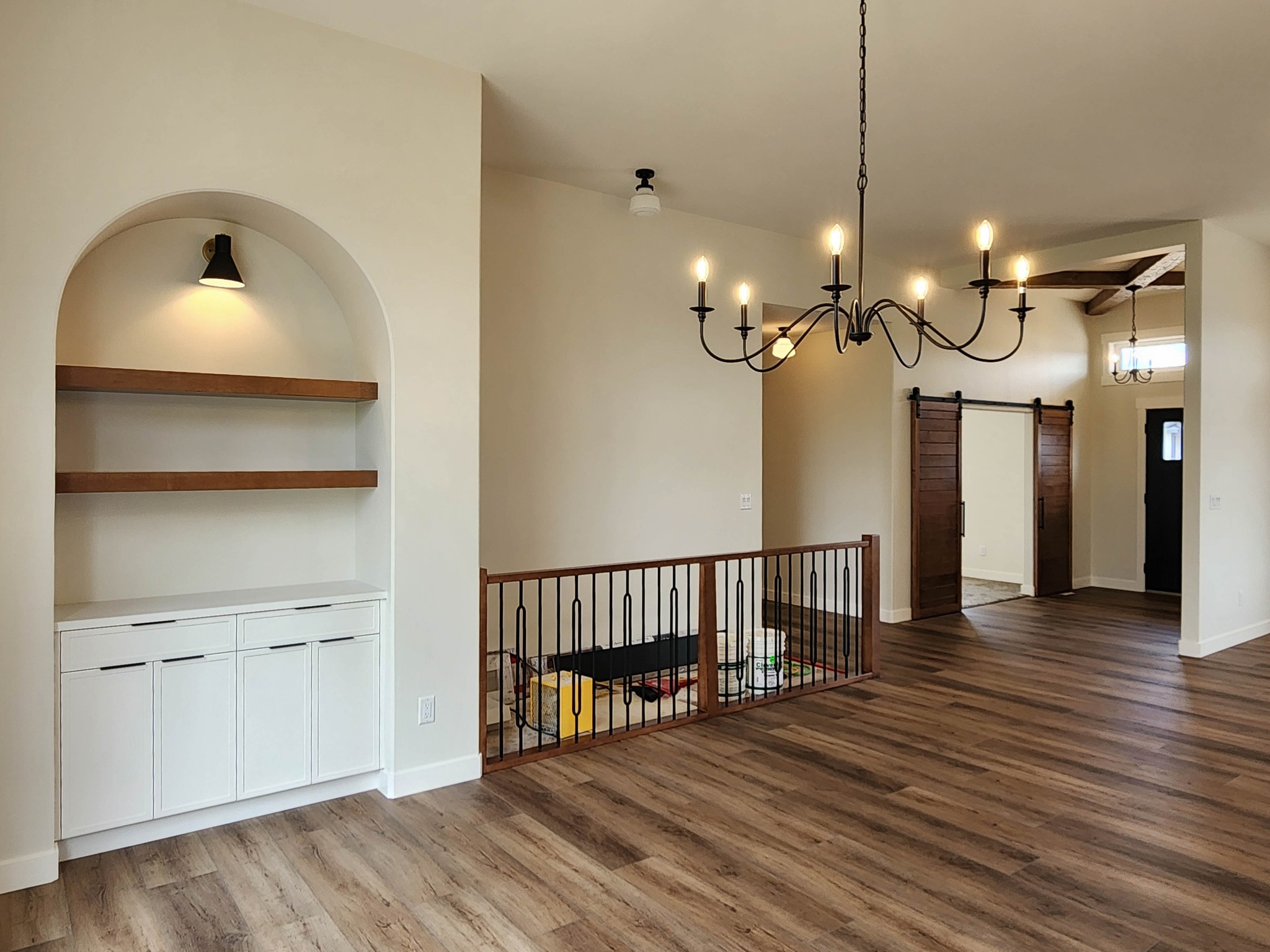 A spacious living area with light wood flooring features a built-in white cabinet with open shelves, black chandelier, sliding barn door, and play area.
