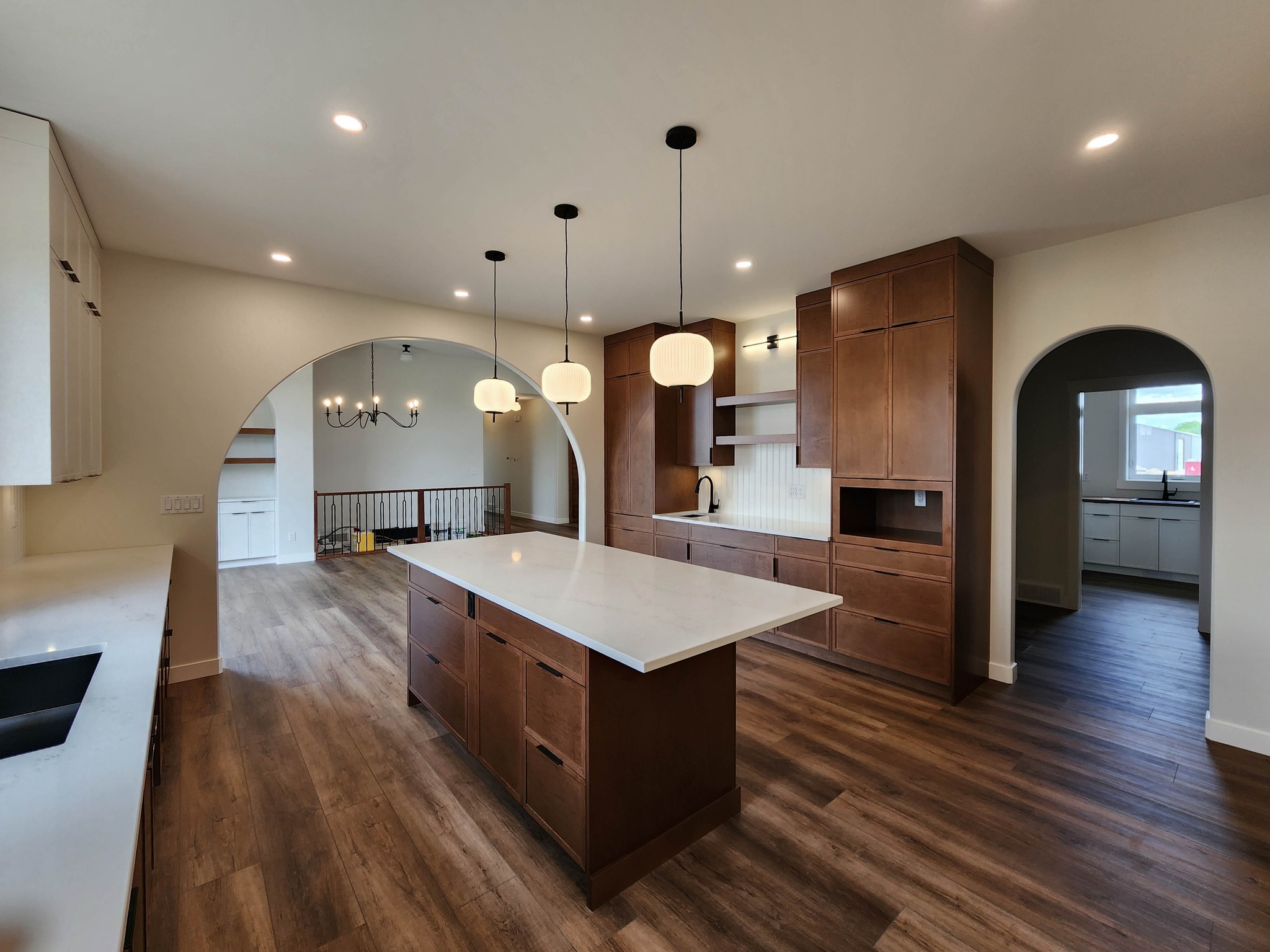 Modern kitchen with wooden cabinets and island, white countertops, and pendant lights. Arched doorways lead to adjacent rooms, creating an open, airy feel.