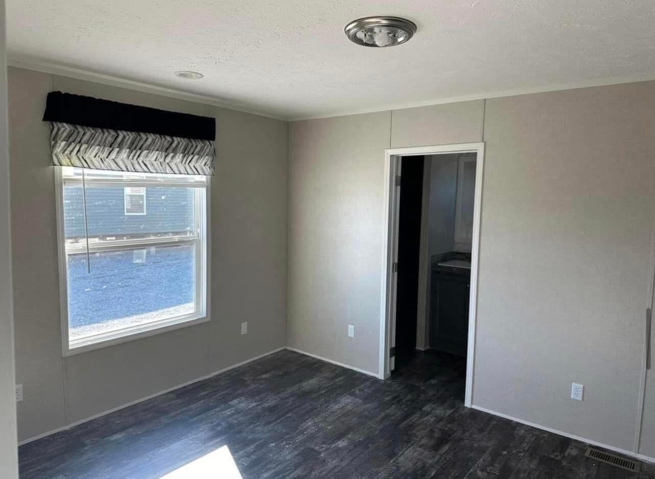 Empty room with gray walls, a large window with a geometric valance, and dark wood flooring. A doorway and ceiling light are visible. Simple, modern feel.