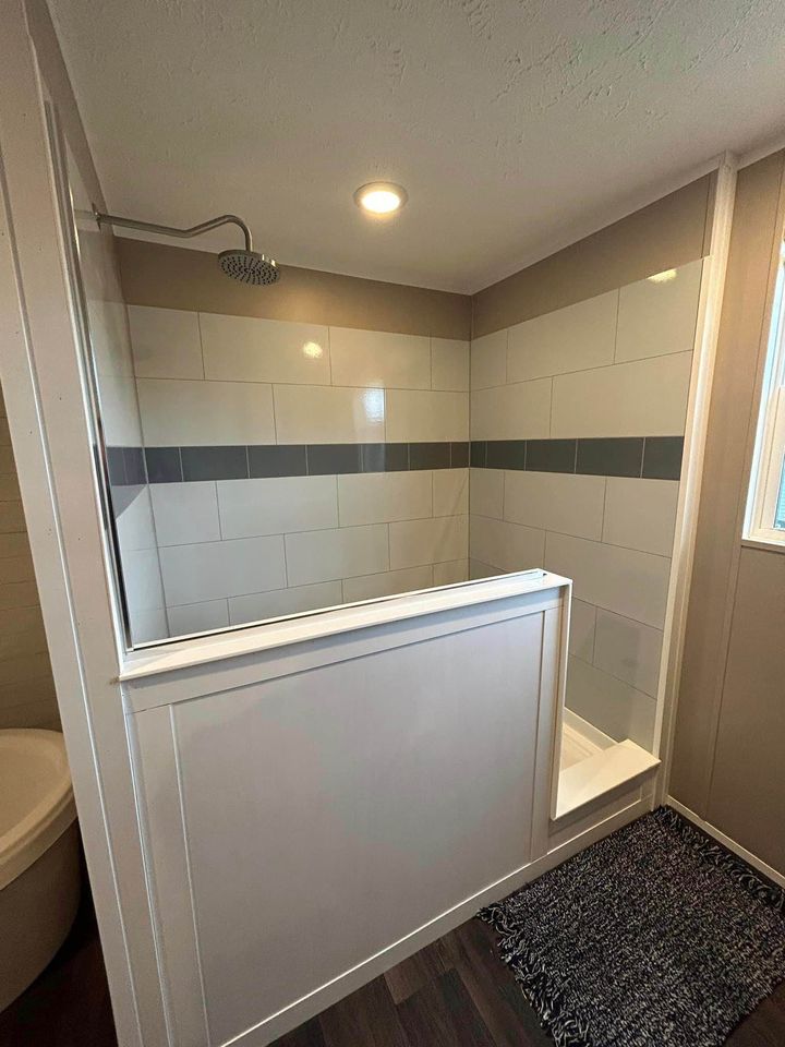 Modern shower with half-wall and glass panel, featuring gray and white subway tiles, overhead rain showerhead, and a textured dark rug on wooden floor.