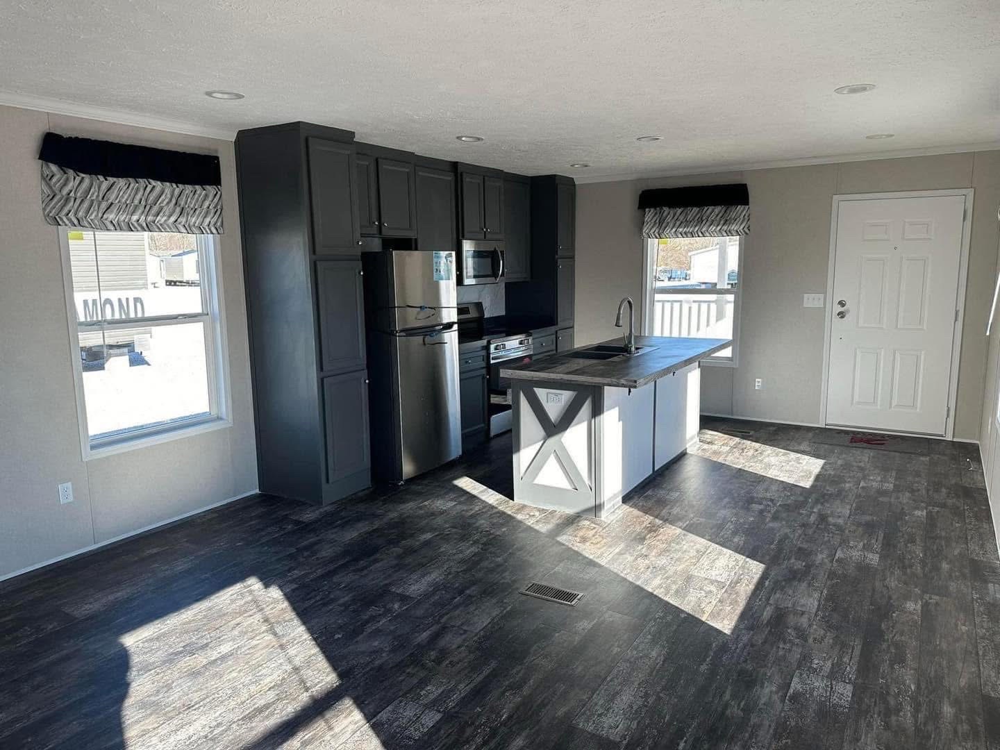 Modern kitchen with dark wood floors, gray cabinets, and stainless steel appliances. A central island with a sink and drying rack is lit by natural sunlight.