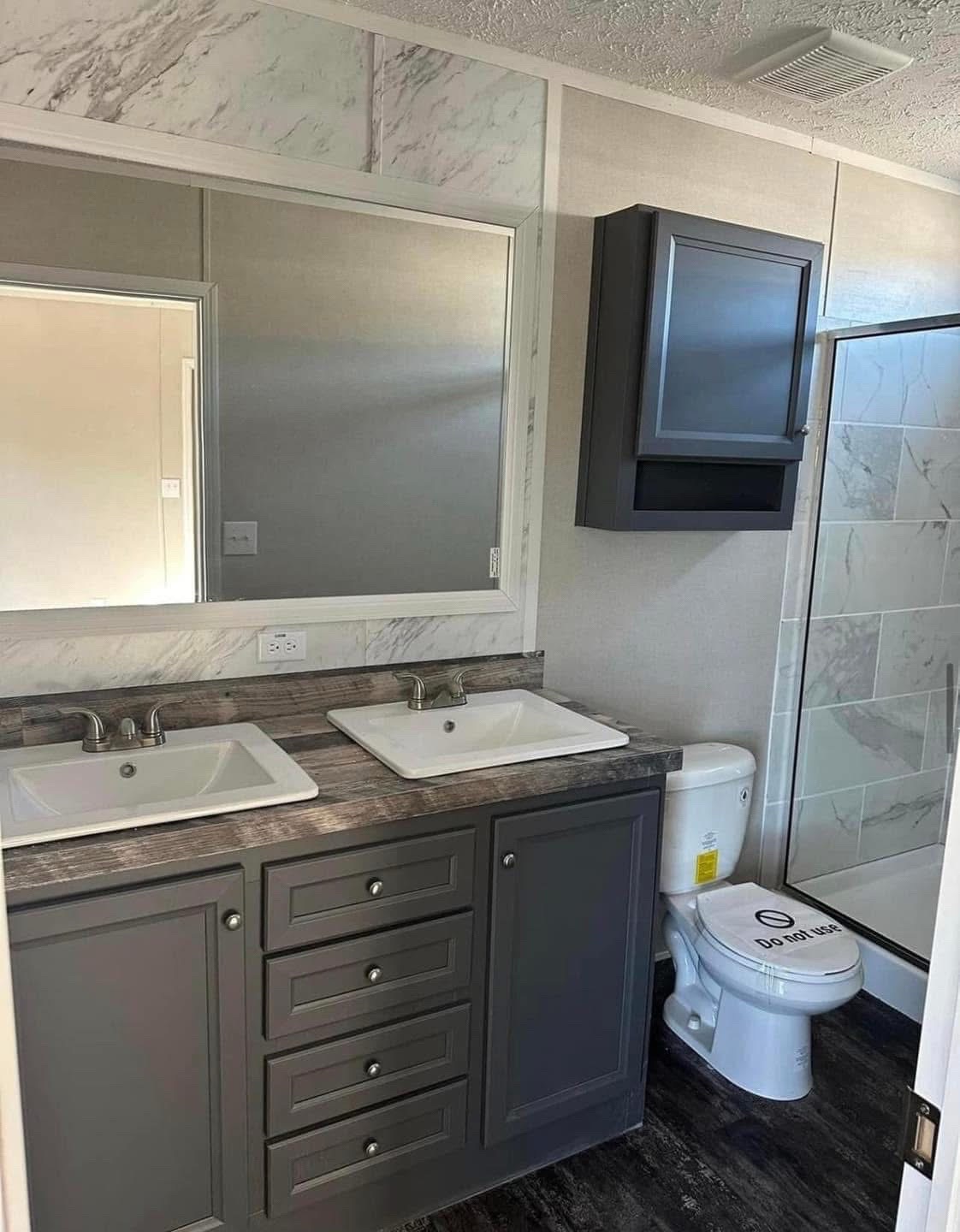 Modern bathroom with dual sinks on a gray vanity, large mirror, and a shower with marble tiles. The toilet has a "Do Not Use" sign.