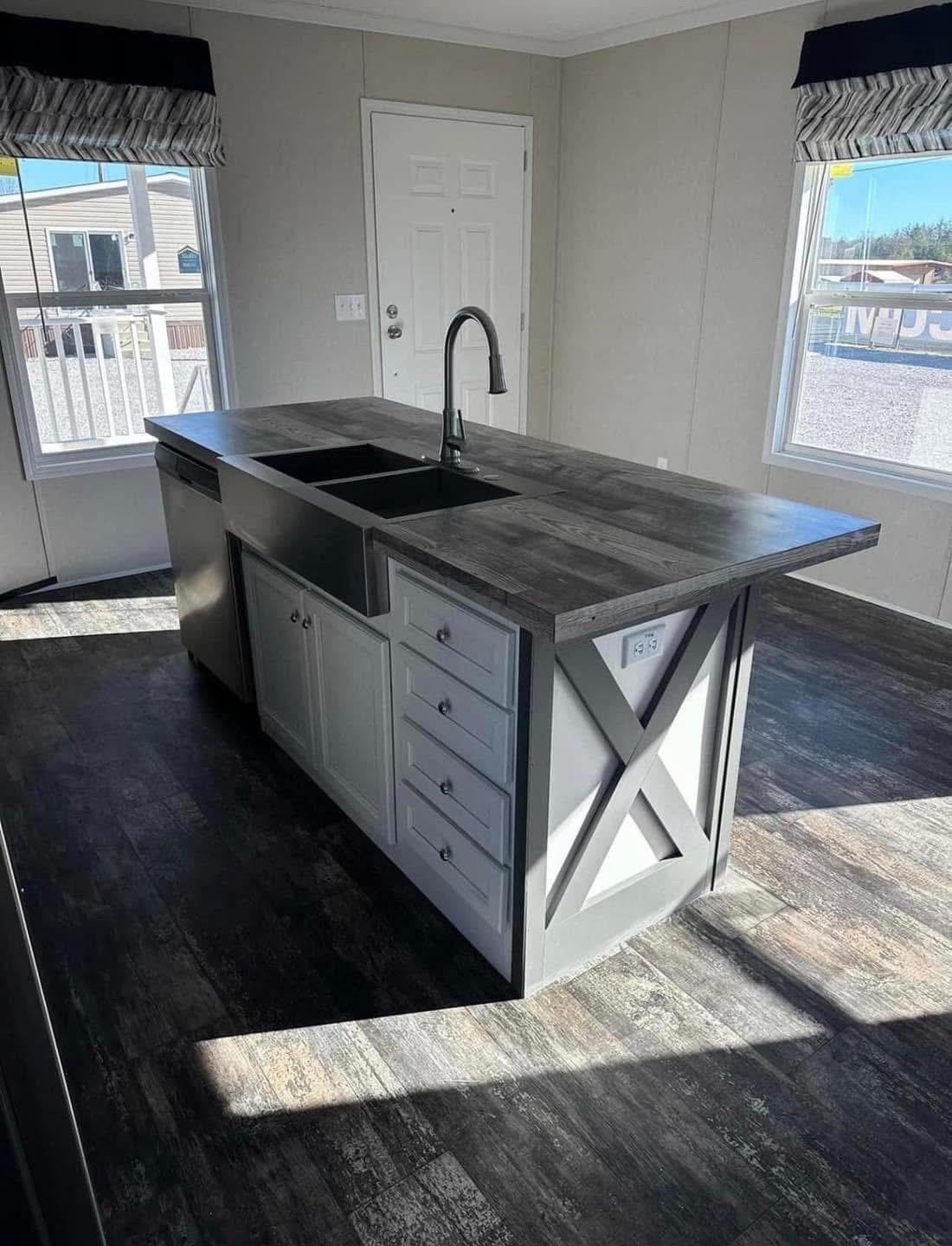 Modern kitchen with a central island featuring dark wood countertops, a black faucet, and white cabinets. Sunlight streams through two large windows.