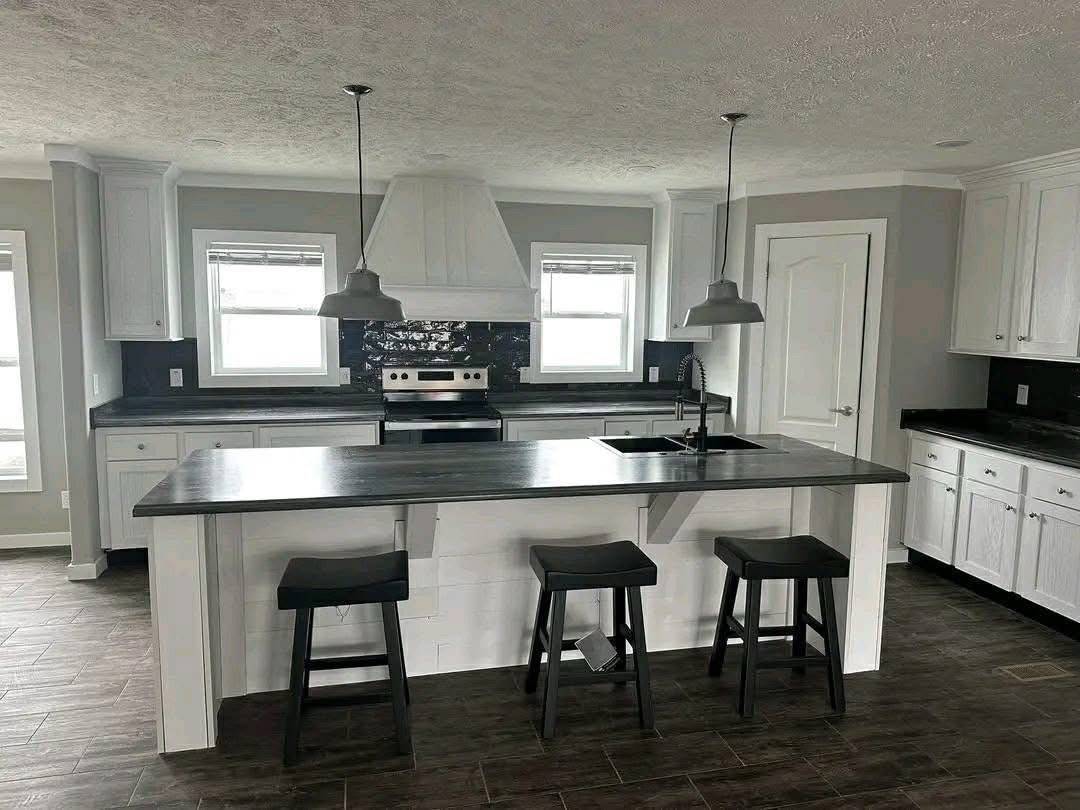 Modern kitchen with dark countertops, a central island with three stools, stainless steel appliances, and white cabinetry. Bright and minimalistic design.