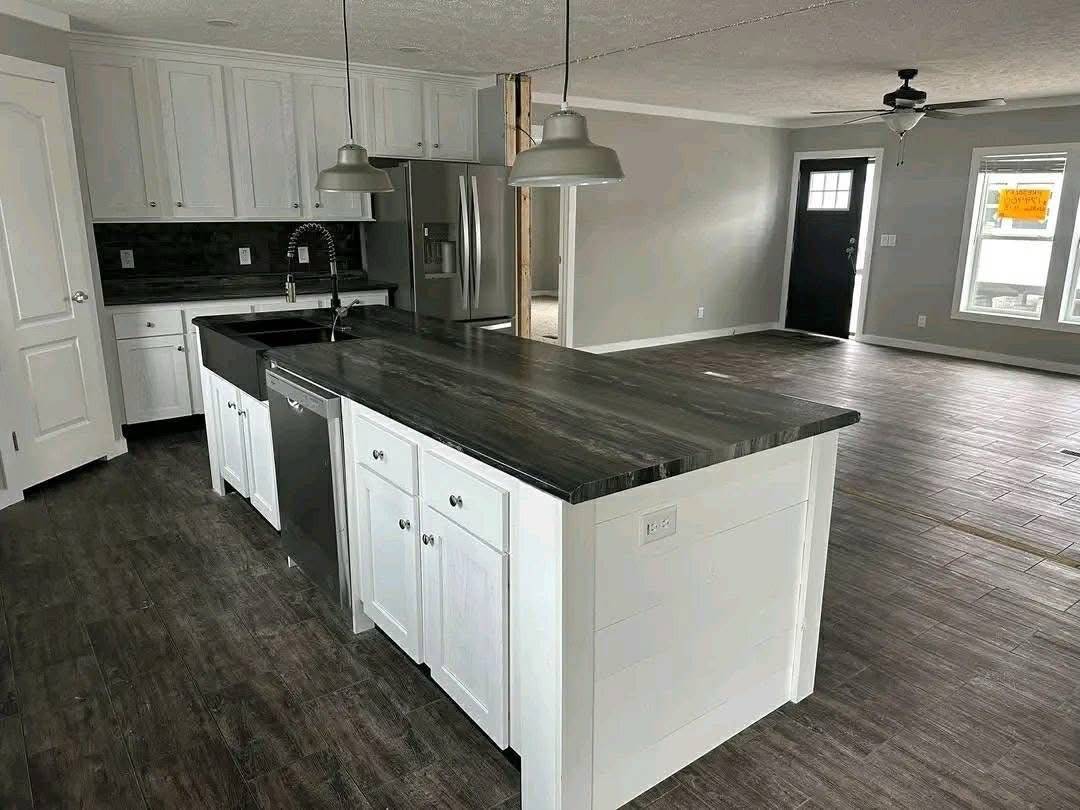 Modern kitchen with white cabinets and black countertops, featuring a large island, stainless steel appliances, pendant lights, and wood flooring.