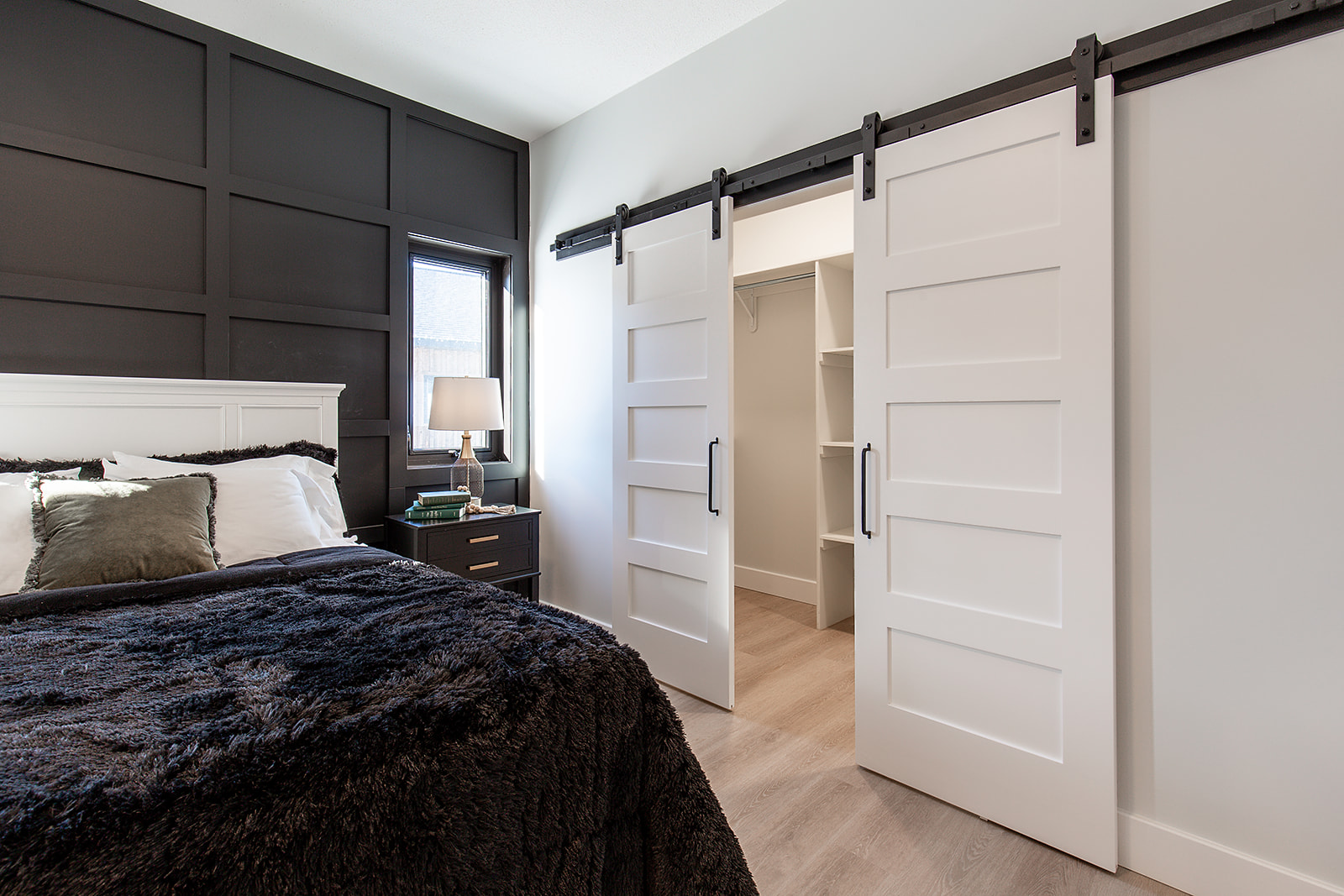 Modern bedroom with dark accent wall, cozy bed, and sliding white barn doors leading to a bright walk-in closet. Soft lighting creates a calm ambiance.