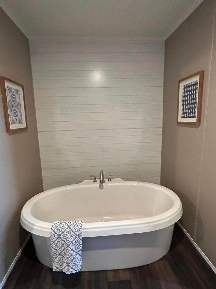 Oval bathtub in cozy bathroom with beige walls, wood flooring, and a shiplap accent. Towel draped over the edge, framed art on walls.