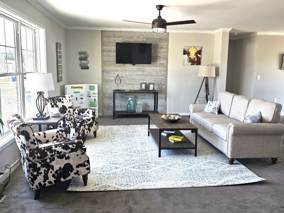 Modern living room with a cozy vibe. Features a beige sofa, cow-print chairs, rustic wooden accents, TV, and a light blue patterned rug, lit by natural light.