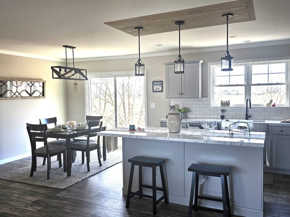 Modern kitchen and dining area with hardwood floors. Features a central island with stools, elegant pendant lighting, and a dining table set by large windows.
