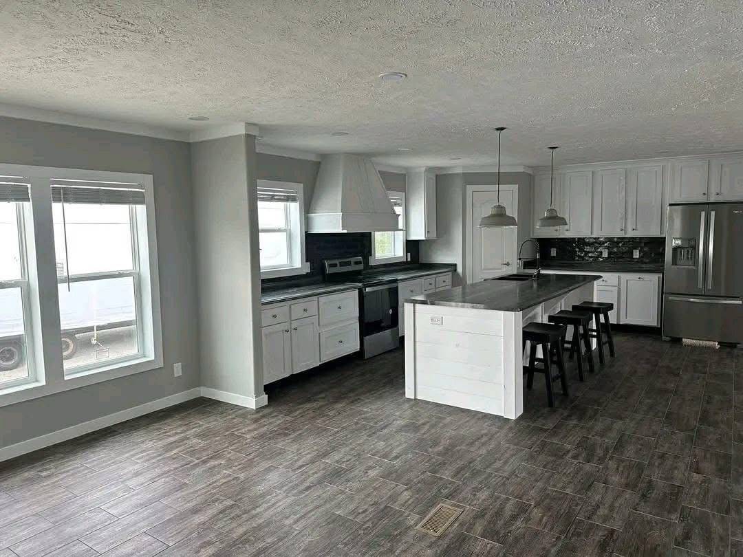 Modern kitchen with white cabinets, gray countertops, and a central island with bar stools. Dark tiled floor, pendant lights, and stainless steel appliances enhance the sleek look. Large windows provide ample natural light.