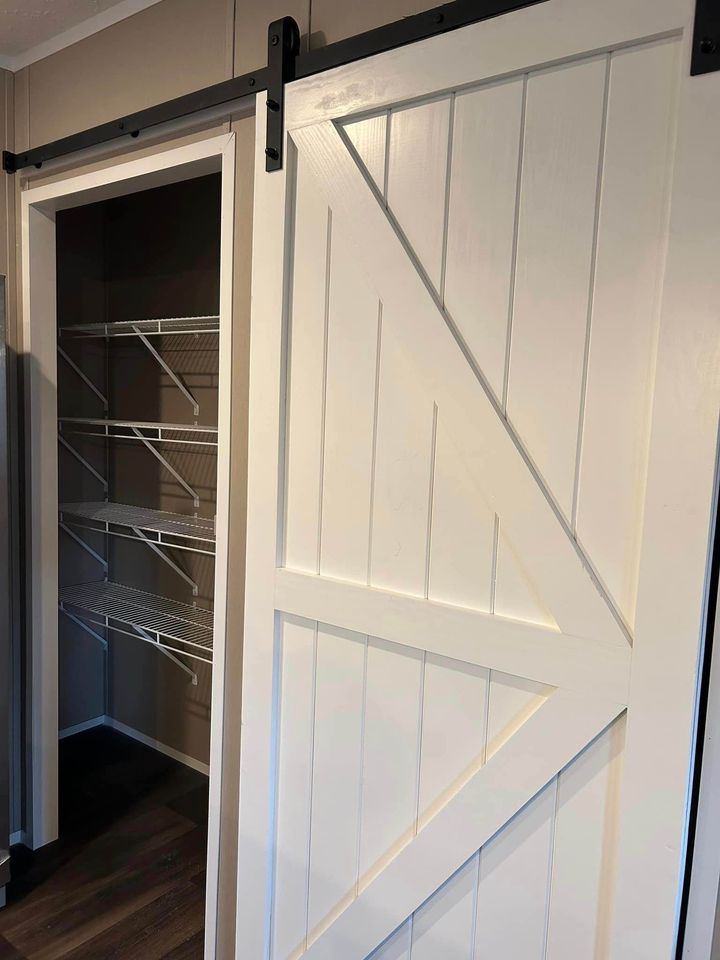 A white sliding barn door is partially open, revealing a closet with empty wire shelves. The room has warm, neutral-colored walls and dark wood floors.