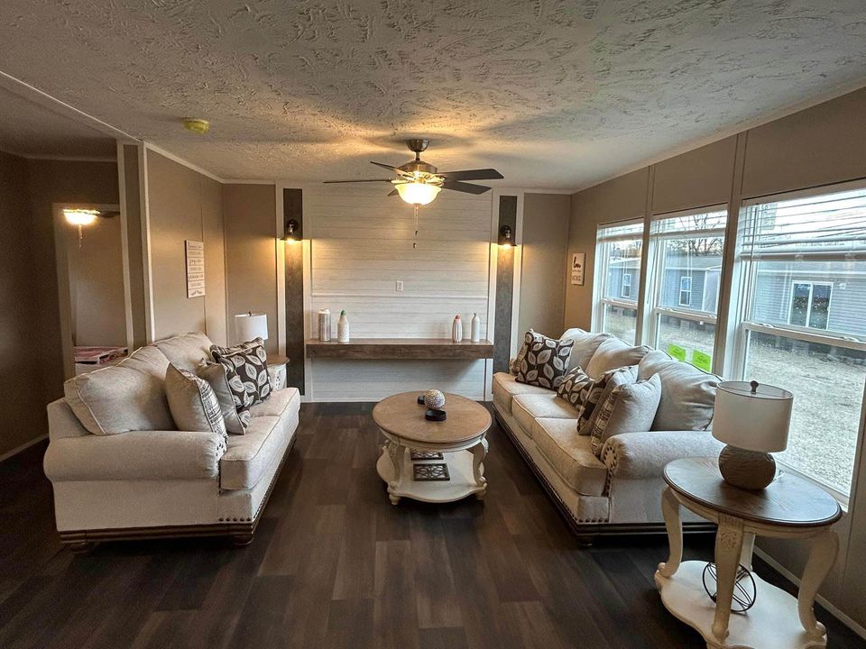 Cozy living room with two beige sofas facing each other, centered around a round wooden coffee table. Large windows on the right provide natural light.