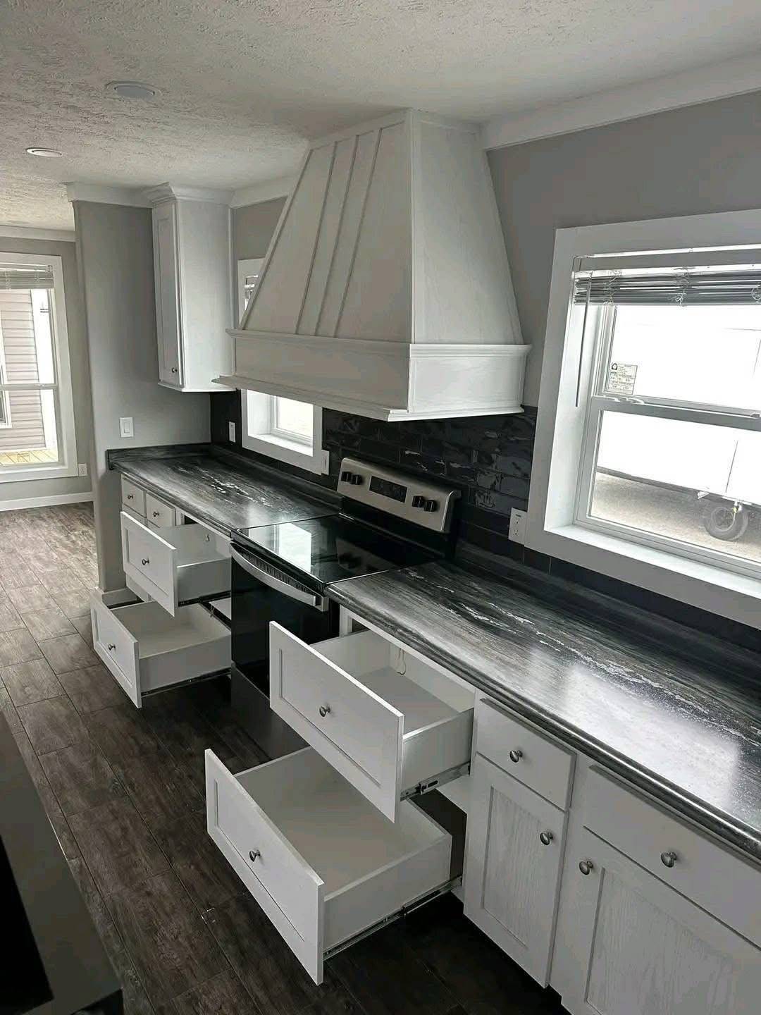 Modern kitchen with white cabinets and drawers open, black countertops, a sleek stove, and two windows. The mood is clean and organized.