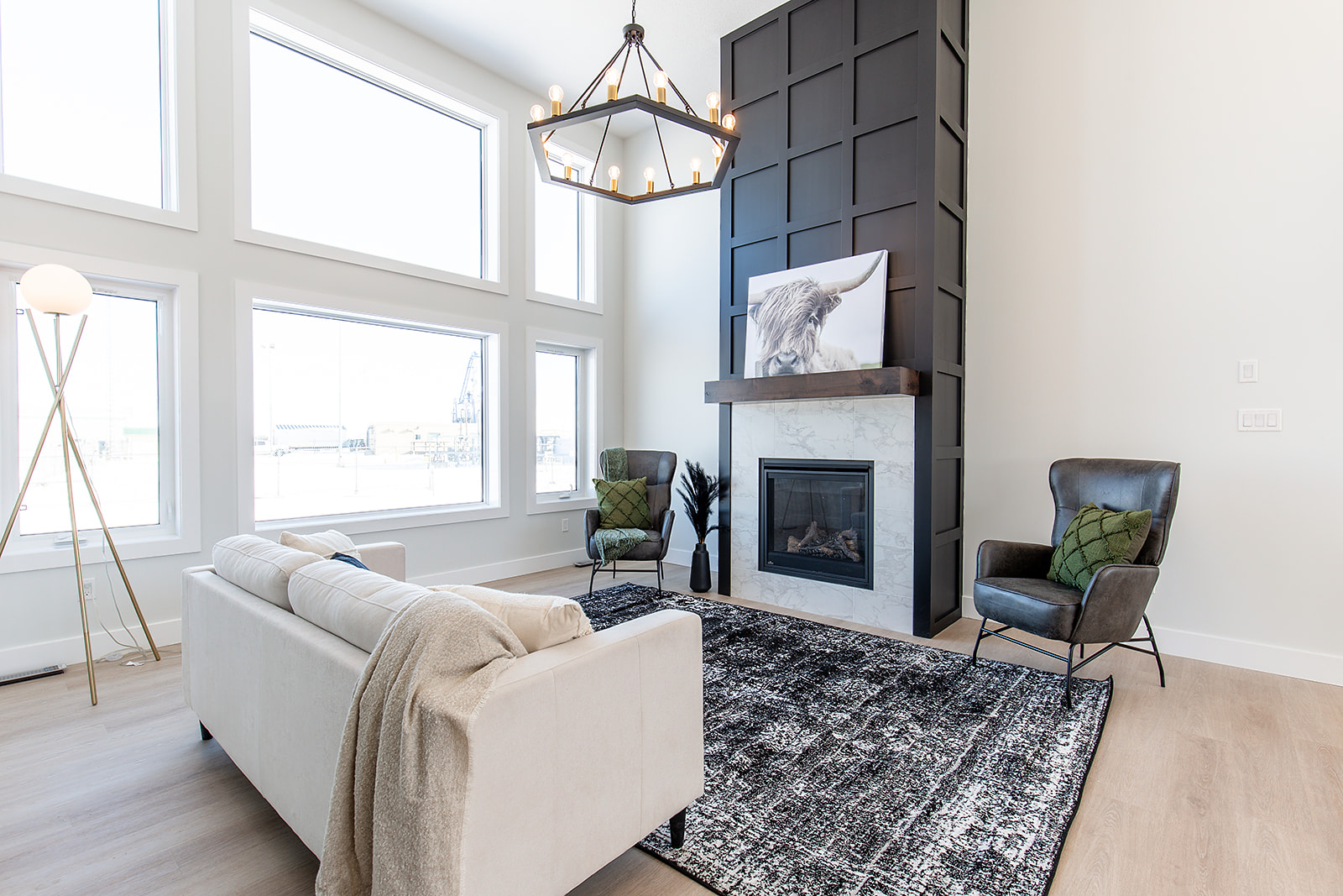 Modern living room with high ceilings, large windows, and natural light. Features a white sofa, black fireplace, and abstract decor with a warm atmosphere.