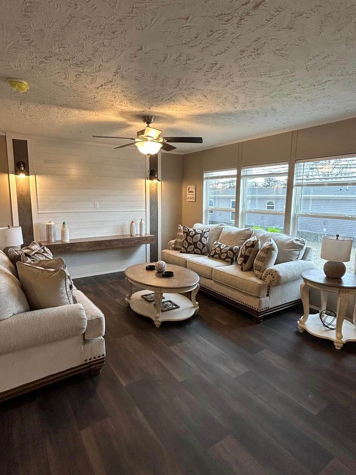 Cozy living room with two beige sofas, patterned pillows, round wooden coffee table, ceiling fan, and large windows. Warm lighting and neutral decor.