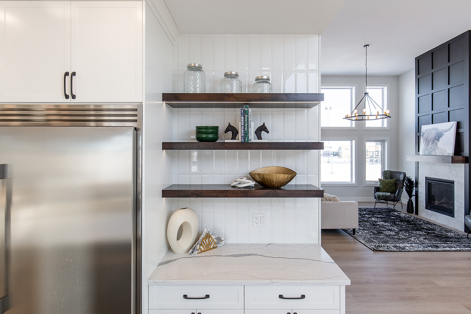 Modern kitchen with wooden shelves displaying glass jars and decor above a white marble counter. Open living area with large windows and fireplace.