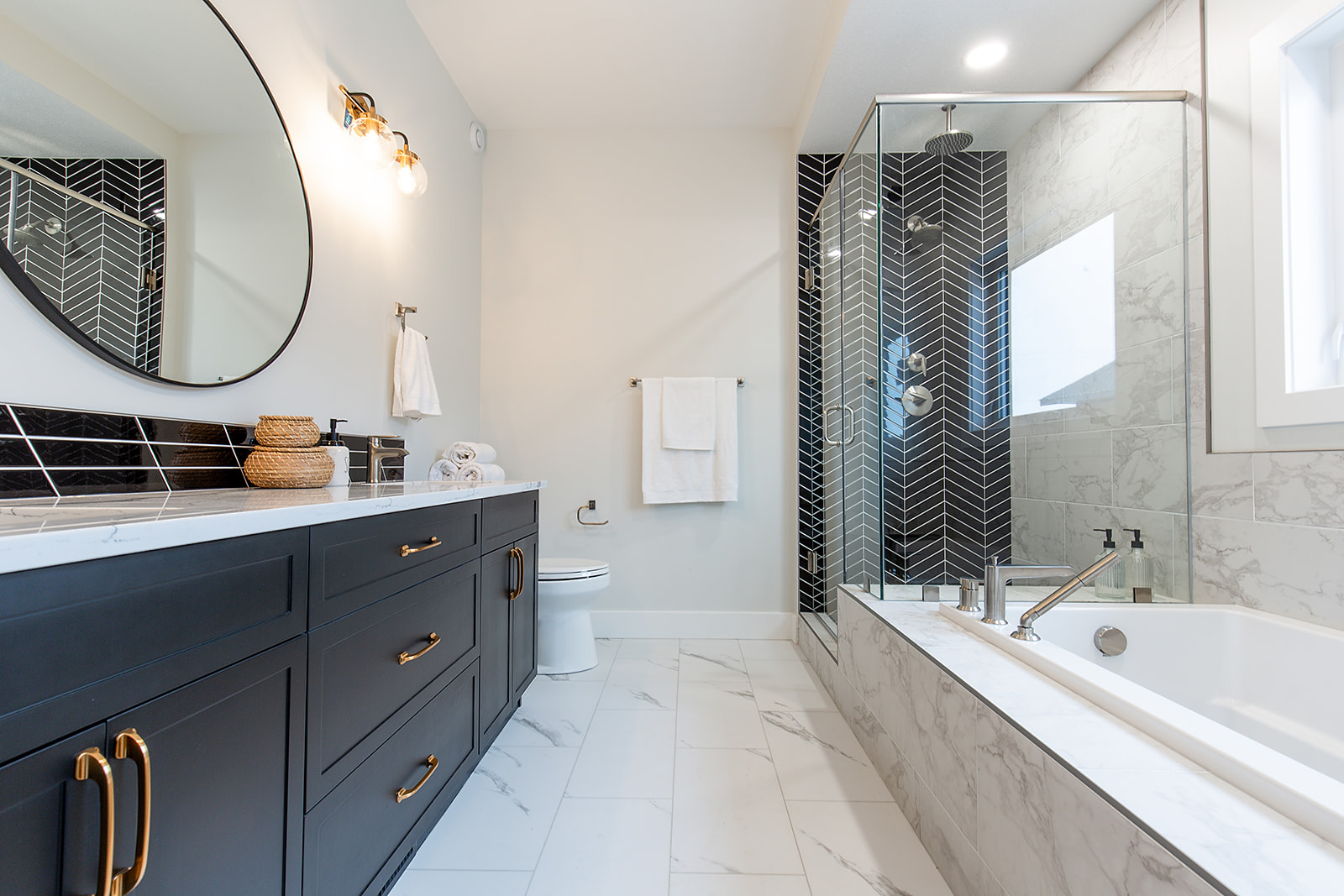 Modern bathroom with a sleek black vanity featuring gold hardware and a round mirror. Glass shower, white marble floor, and a large tub create an elegant feel.