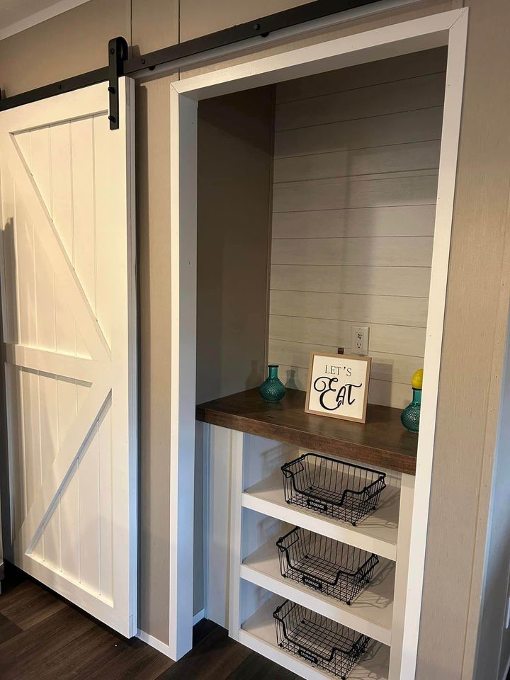 A white sliding barn door next to a pantry nook with shelves. The shelves hold wire baskets and a wooden counter features vases and a "Let's Eat" sign.