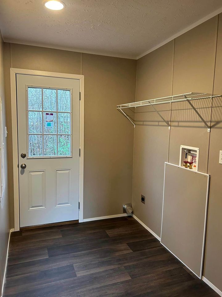 A small, empty laundry room with beige walls, a wooden floor, a white door with glass panes, and a wire shelf. The space feels clean and minimalistic.