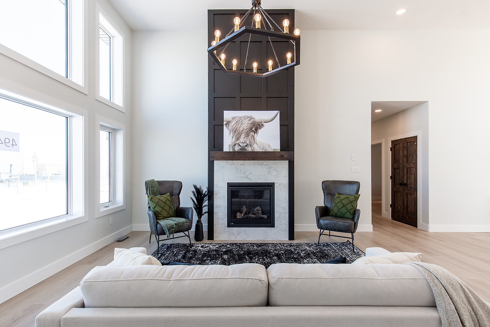 Modern living room with high ceiling, chandelier, and large windows. Features a central fireplace with animal artwork, flanked by two chairs, and a sofa.