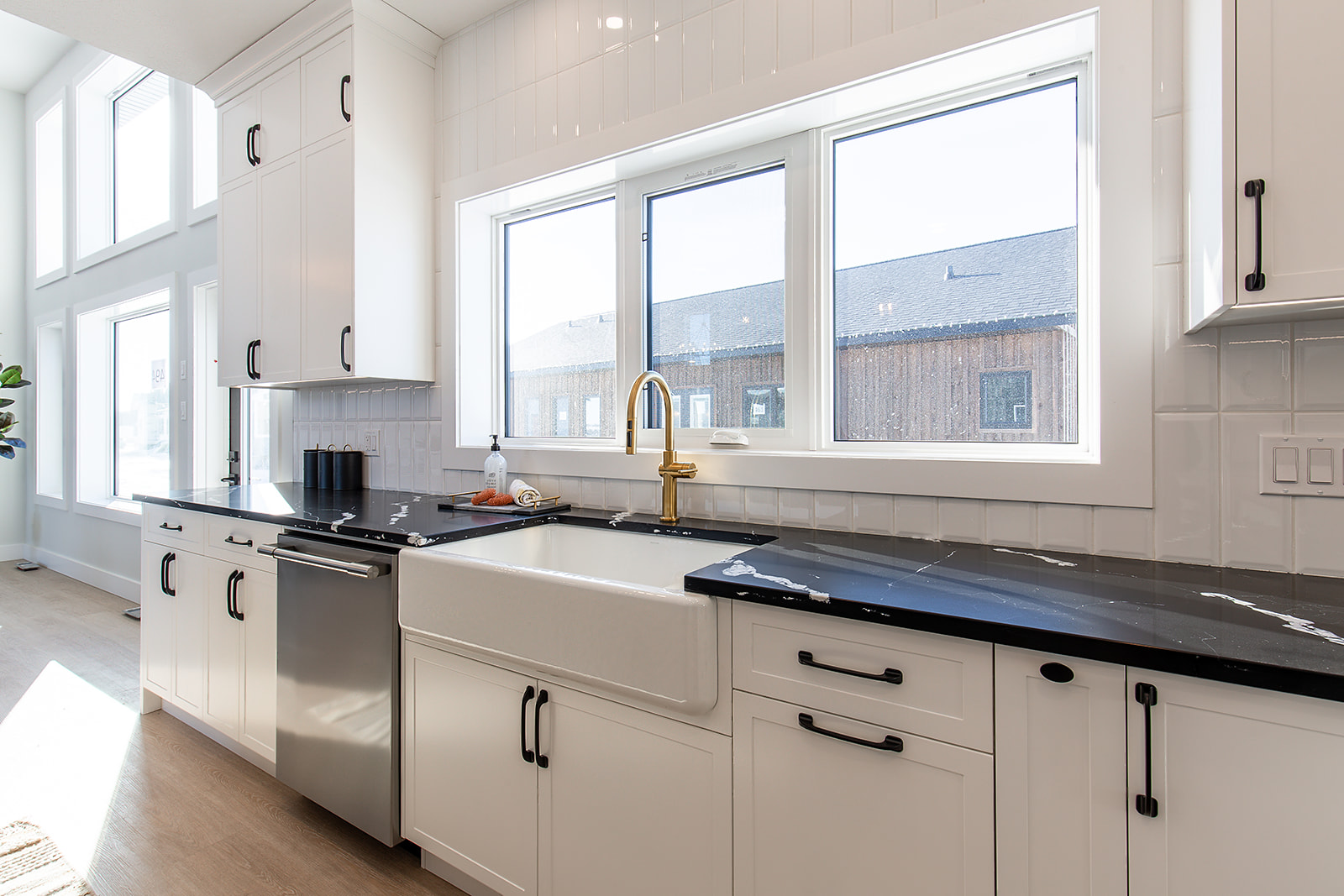 Bright modern kitchen with white cabinetry, black countertops, and a gold faucet over a farmhouse sink. Large windows flood the space with light.