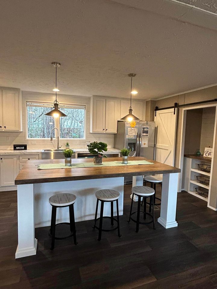Modern kitchen with a wooden island and four stools, white cabinets, stainless steel appliances, hanging lights, a large window, and a barn-style pantry door.