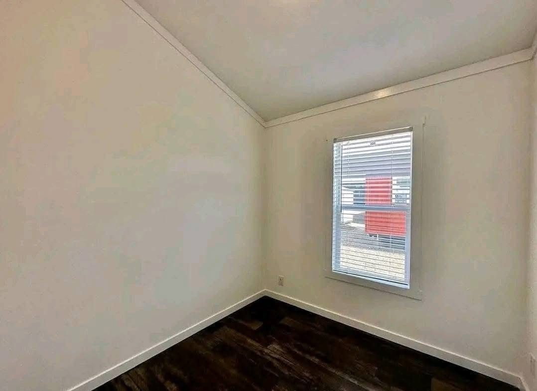 A small, empty room with light cream walls, a sloped ceiling, and a single window with blinds. The dark wood floor contrasts with the bright space.