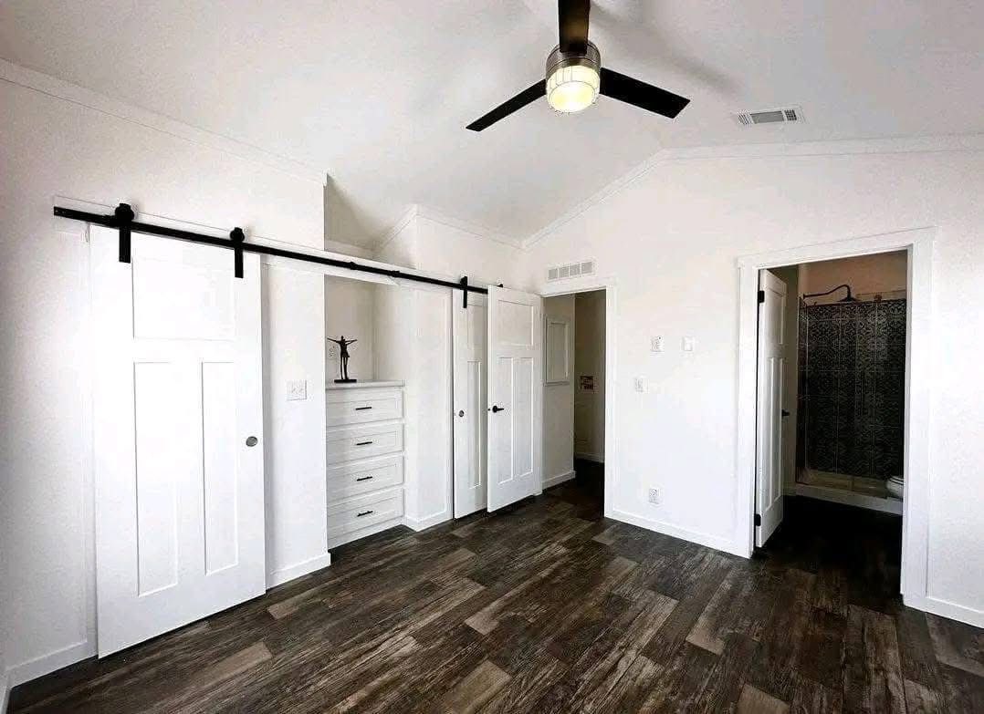 Minimalist white room with wooden floors, featuring sliding barn doors, a black ceiling fan, and an open doorway revealing a bathroom.