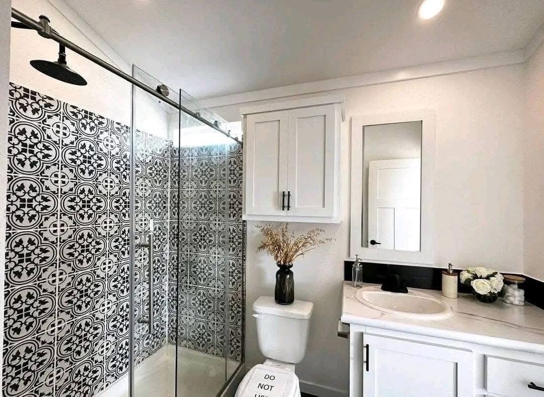 A modern bathroom features a black and white patterned tile shower, glass doors, and a rain showerhead. A white cabinet, vase with dried flowers, and white sink complete the sleek look.