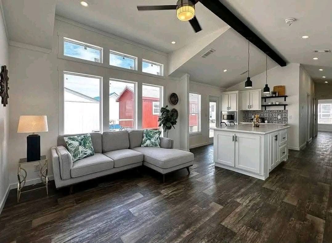 Modern living room with high ceilings, featuring gray sectional sofa, tropical pillows, and lamp. Adjacent kitchen boasts white cabinets and island. Bright and airy.