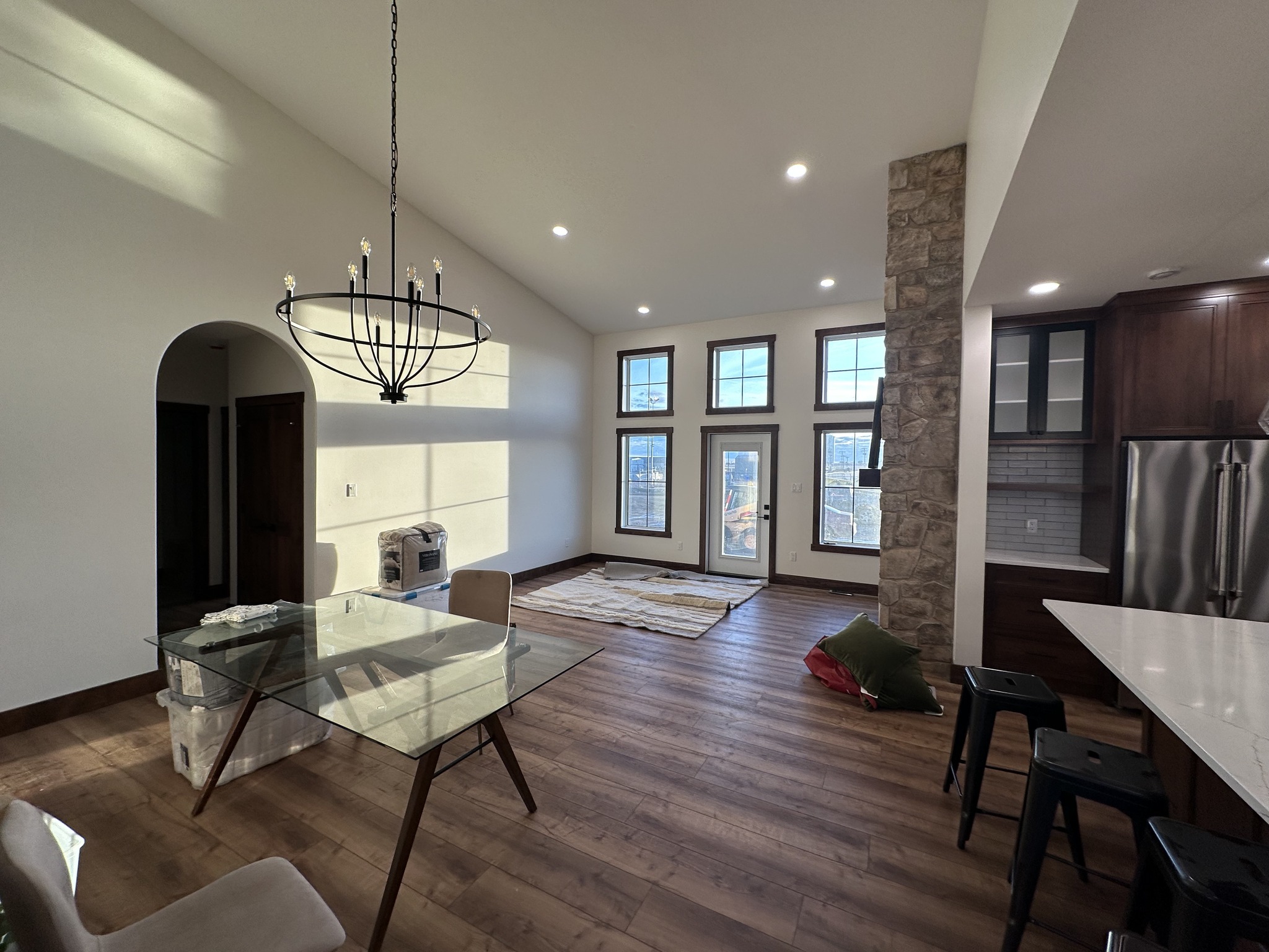 A modern living space with high ceilings and warm wood floors. Features a glass dining table, large windows, a stone accent wall, and a stylish chandelier.