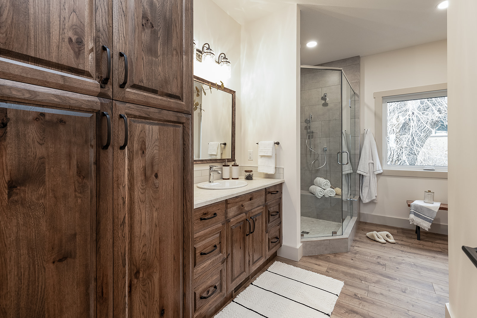 Spacious bathroom with rustic wooden cabinets, dual sinks, and a glass shower. Soft, natural light from a window creates a cozy, inviting atmosphere.