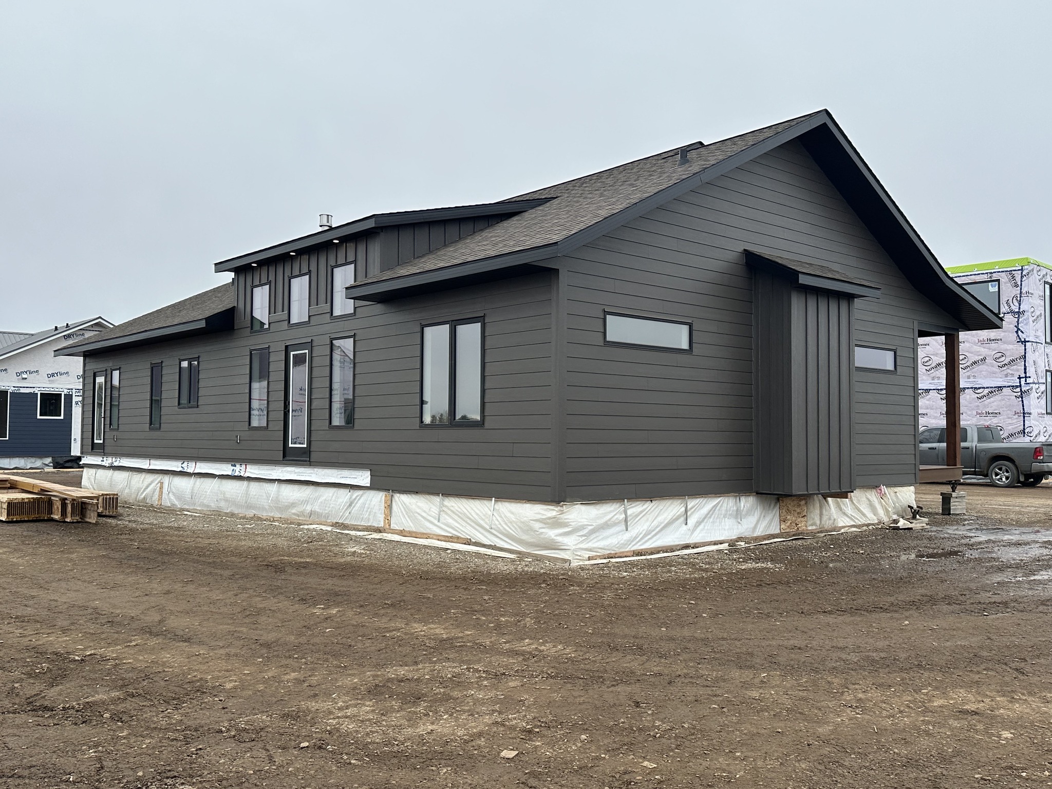 Gray modular home under construction on a muddy lot, wrapped in protective sheeting. Overcast sky, adjacent buildings visible in the background.