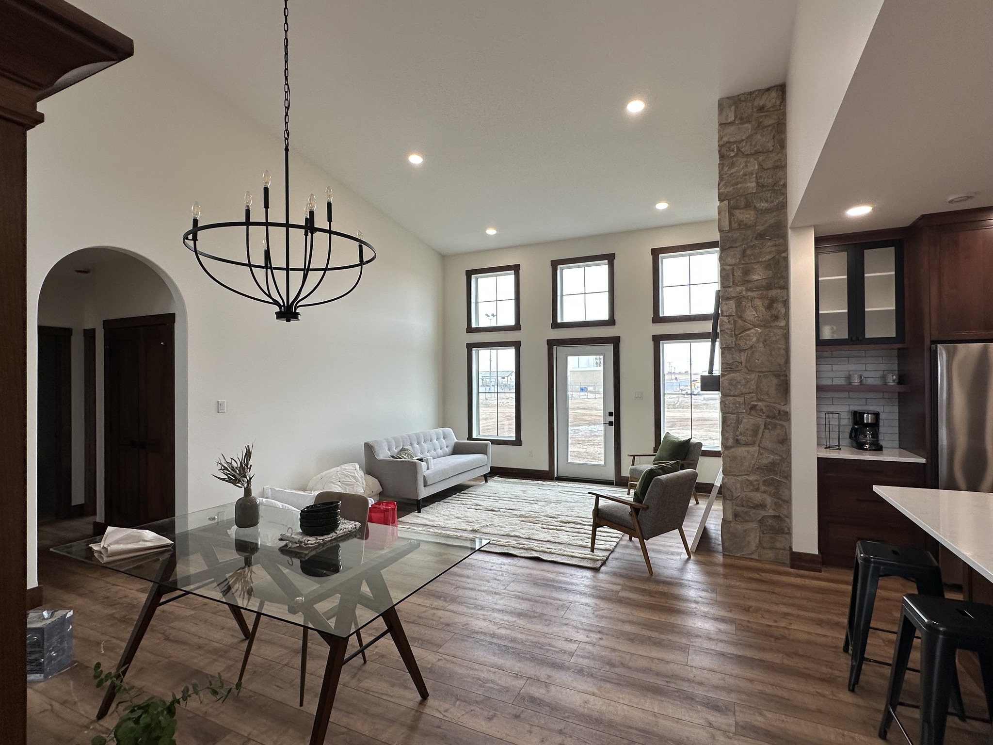 Modern living room with high ceiling and large windows. Features a gray sofa, glass dining table, stone accent wall, wooden flooring, and chandelier. Bright, airy ambiance.