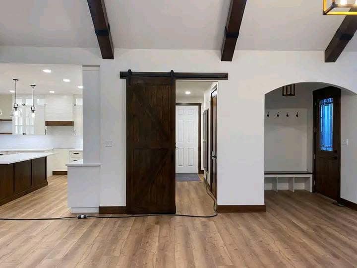 Modern interior with wooden floors and beams, featuring a dark sliding barn door. To the left is a white and wood-accented kitchen; to the right, an entryway with coat hooks.