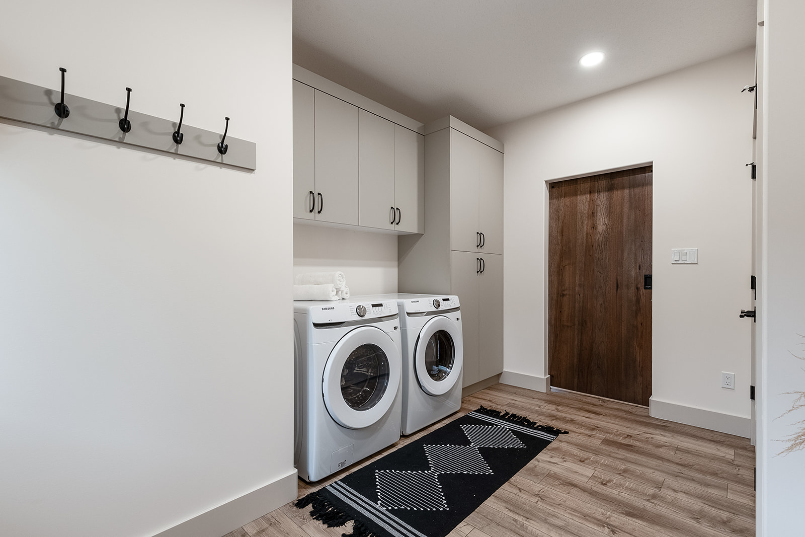 Modern laundry room with light wood flooring, featuring a white washer and dryer. Gray cabinets above and to the side. A dark rug adds contrast.