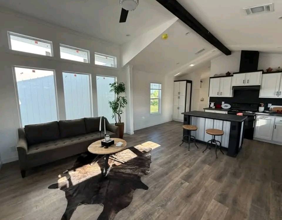 Modern living room and kitchen with high ceilings and dark wood beams. It features a gray sofa, round table, cowhide rug, and an island with stools. Bright and airy atmosphere.