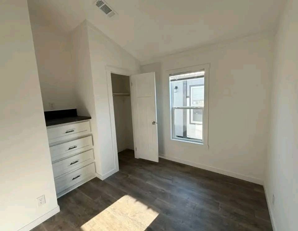 A small, empty room with white walls and a wooden floor. There's a set of closed drawers and an open closet on the left, and a window on the right letting in sunlight.