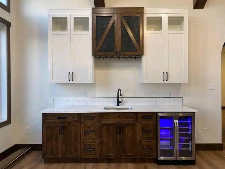 Modern home bar with white upper cabinets and dark wooden lower cabinets. Features a sink, wine rack, and fridge with blue interior light.