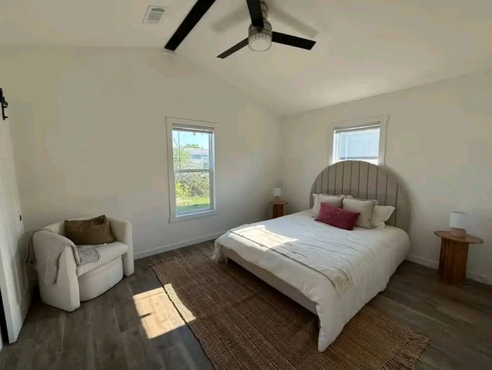 A minimalist bedroom with a large bed featuring white linens and a curved headboard. There's a cozy armchair, a woven rug, and two wooden side tables.