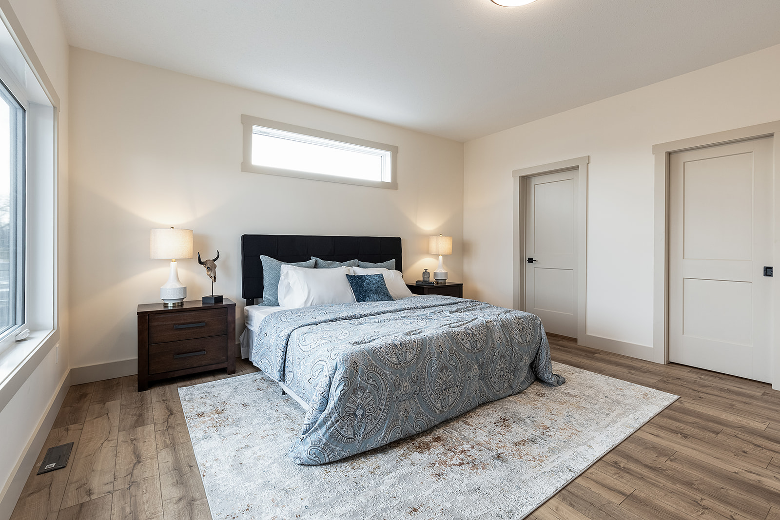 A cozy bedroom with a large bed featuring a patterned blue bedspread and numerous pillows. Wooden nightstands with lamps flank the bed, creating a warm and inviting atmosphere. Sunlight filters through a high horizontal window, and the room has light walls, wooden floors, and a soft rug, conveying a serene and modern style.