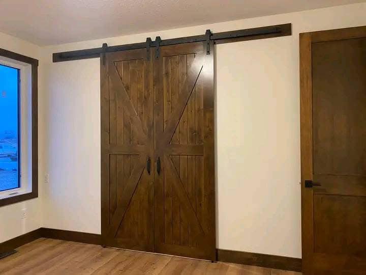 A room with wooden barn-style sliding doors on the wall, adjacent to a regular door. Large window on the left, wood flooring, light walls. Cozy ambiance.