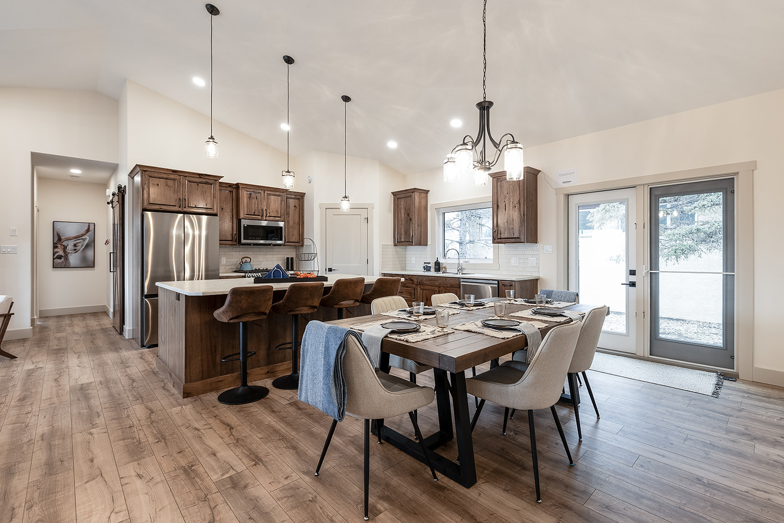 Modern kitchen and dining area with rustic charm, featuring wooden cabinets and floors. A large table is set for six under elegant pendant lighting.
