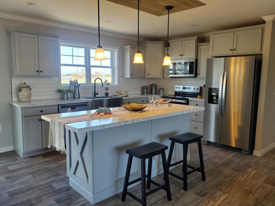 Modern kitchen with light gray cabinets, marble island, and pendant lights. Stainless steel appliances and two black stools add contrast to the warm ambiance.