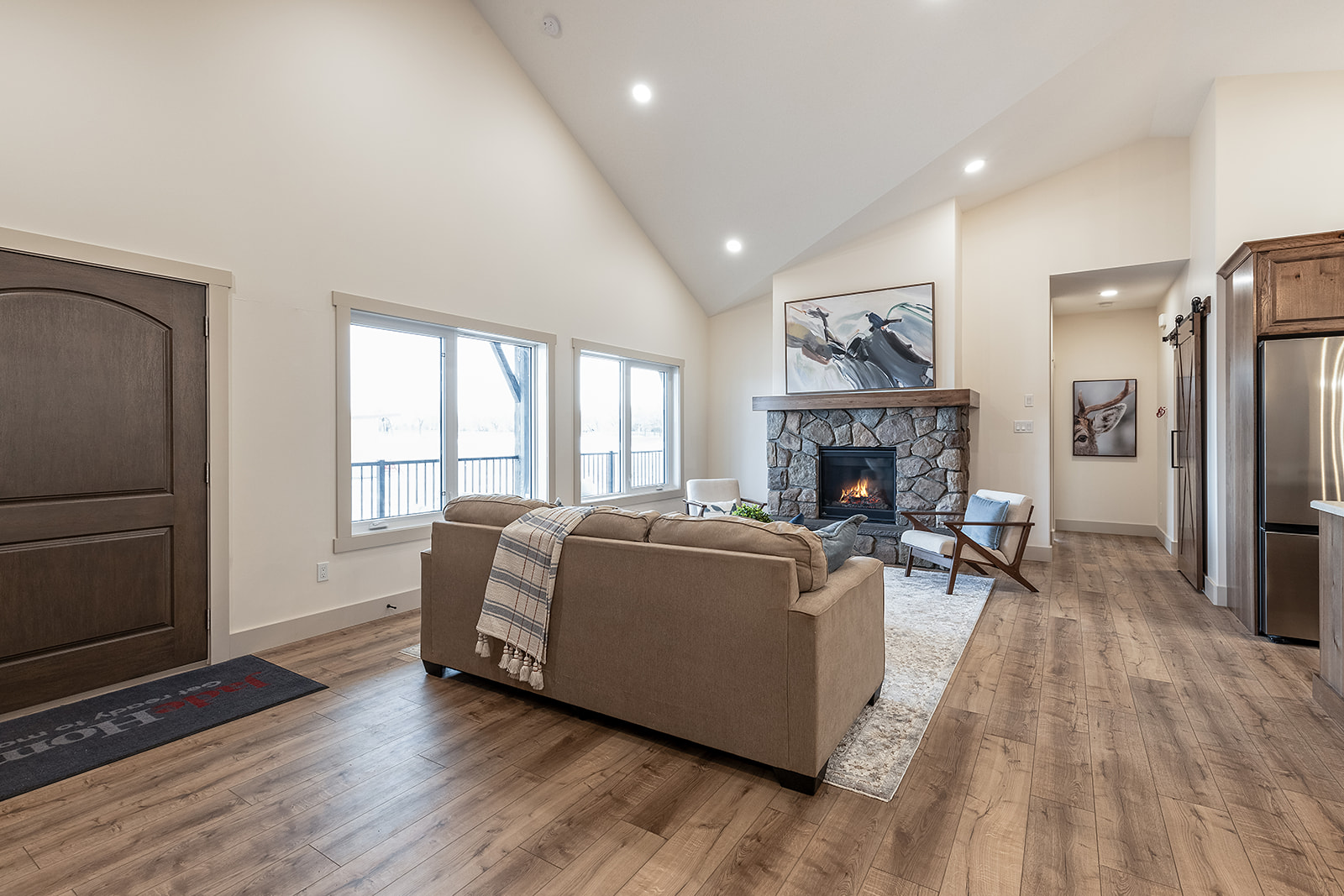 Spacious living room with vaulted ceilings, featuring a cozy stone fireplace, a beige sofa with a plaid throw, and warm wood flooring. The room is well-lit by large windows, creating an inviting and comfortable atmosphere.