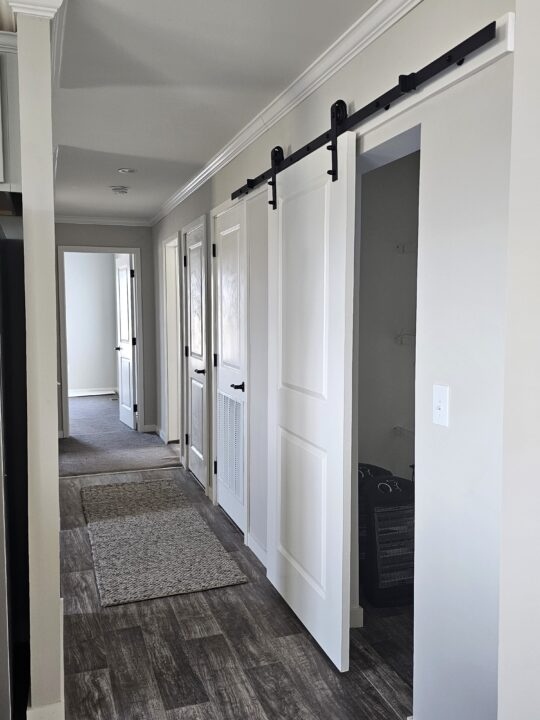 A hallway with wooden flooring features white barn doors on black sliding tracks. Neutral-tone walls and soft lighting create a calm, modern ambiance.