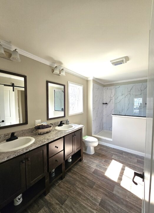 Spacious bathroom with dual sinks and a large mirror above each. Marble-patterned shower in the corner, wooden floors, and bright natural lighting.
