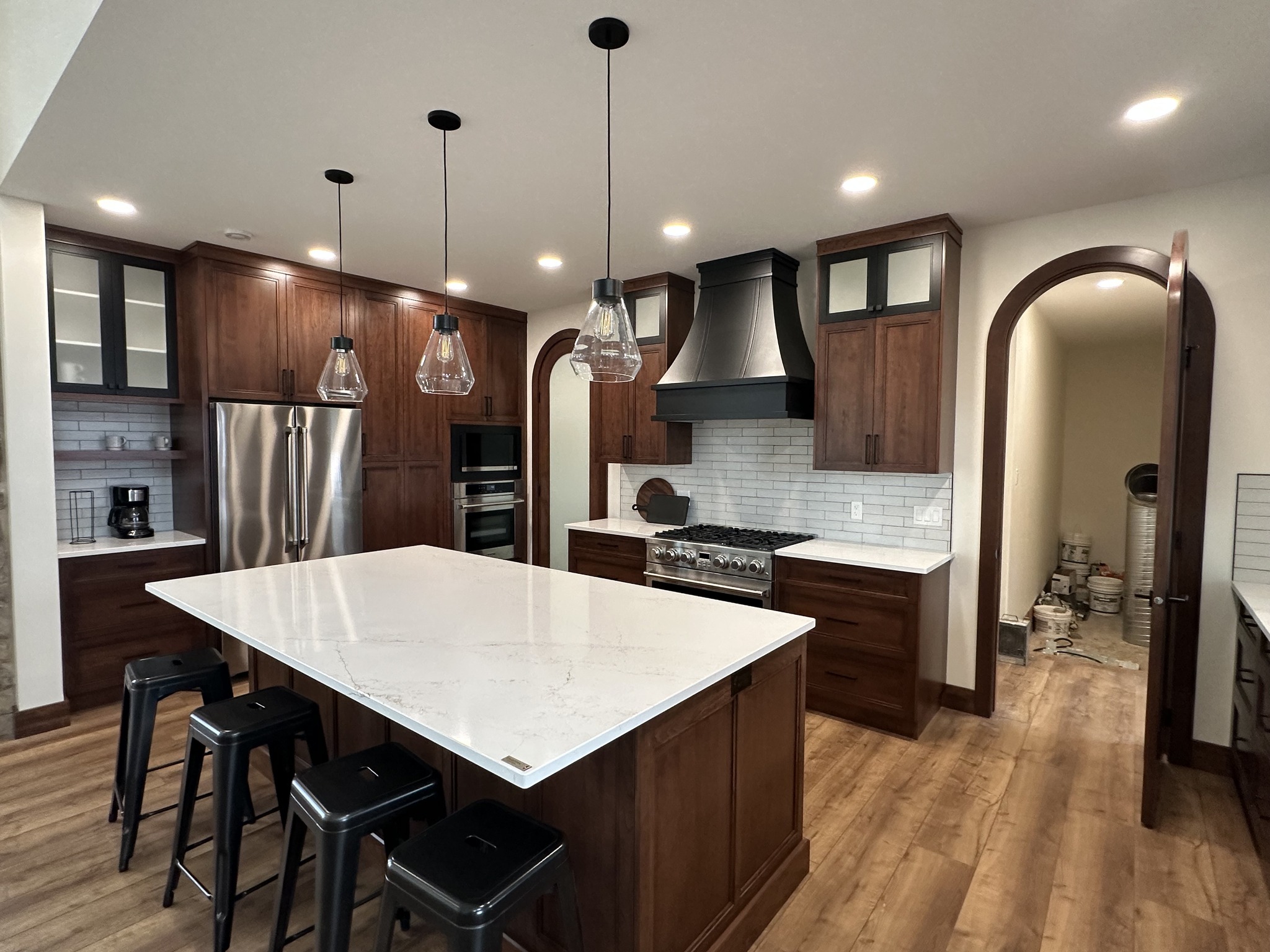 Modern kitchen with wooden cabinets, white countertops, and hardwood floors. Features an island with black stools, pendant lights, and stainless steel appliances.